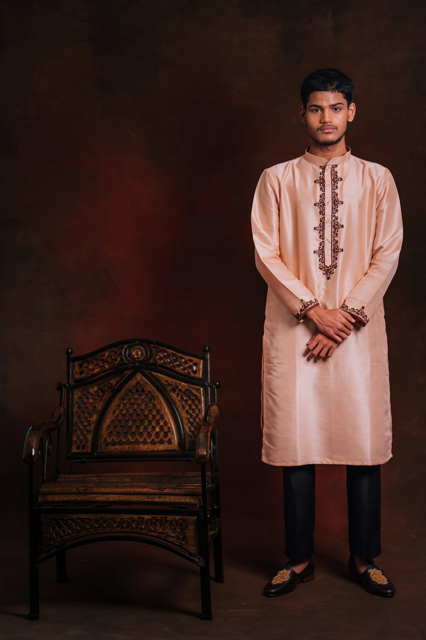 Model wearing a light pink kurta with intricate embroidery by team AKloset standing next to a wooden chair against a dark background.